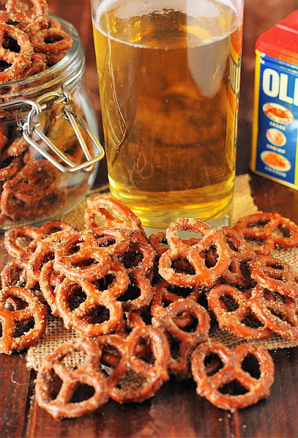 A rustic setting featuring seasoned mini pretzels with a visible coating of spices, scattered on burlap cloth. In the background, a glass jar filled with more of these pretzels sits open, next to a tall glass of beer with foam on top, suggesting a pairing for a casual snack. To the right, there is a box of Old Bay seasoning, hinting at the flavor profile of these crunchy treats.