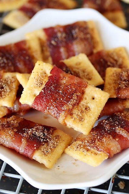 An array of sweet and spicy bacon crackers cooling on a wire rack. Each cracker is wrapped with a strip of bacon and generously sprinkled with a dark brown sugar and chili spice mixture, giving them a caramelized appearance. The seasoned bacon provides a stark contrast to the golden hue of the buttery crackers beneath.